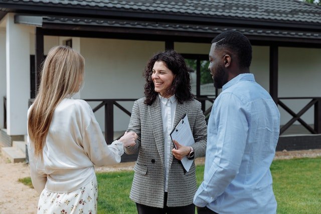 Three People Standing In Front Of A House, Two Of Them Are Shaking Hands