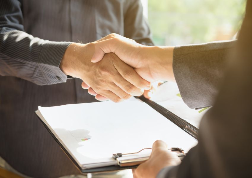 Two People Wear Dark Colored Suits Shaking Hands Across A Desk With A Document On It