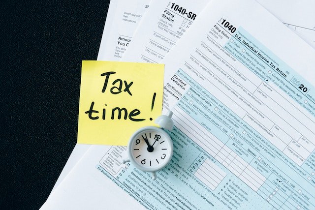 Small Clock, Post-It Note Reading“Tax Time,” And Tax Documents On A Black Table